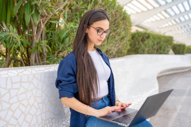 Gözlüklü, parktaki bankta oturan ve dizüstü bilgisayar kullanan esmer kadının portresi.