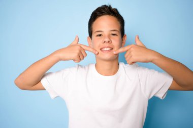 Dişlerini gösteren bir gencin portresi. Parmak dişleri ve ağzı olan bir çocuk. Kafkasyalı genç, mavi arka planda izole..