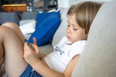 Kulaklıklı çocuk evde akıllı telefon kullanarak koltukta oturuyor.