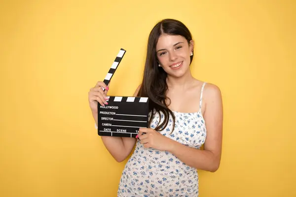 stock image Beautiful cheerful happy young brunette girl teen student wears summer dress holding classic black film making clapperboard isolated on yellow background