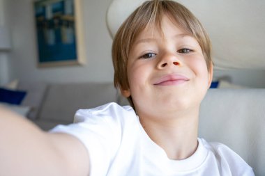 Sevimli beyaz çocuk evdeki kanepede oturmuş kamerayla selfie çekiyor.
