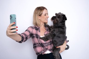 Genç gülümseyen bir kadın küçük köpeğine sarılıyor birlikte selfie çekiyor ve beyaz arka planda duruyor..