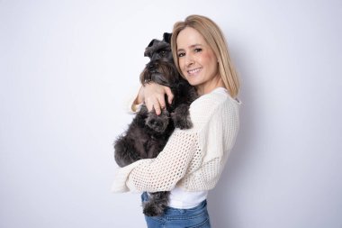 Genç sarışın kadın gülümsüyor köpeğine sarılıyor beyaz arka planda tek başına duruyor..