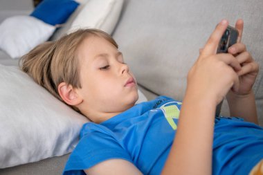 Bir kanepede uzanmış akıllı telefon kullanan mavi tişört giyen ciddi bir çocuğun portresi. Teknoloji konsepti.