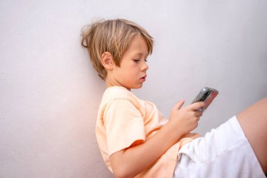Beyaz duvar arkasında akıllı telefon kullanan sarışın çocuk..