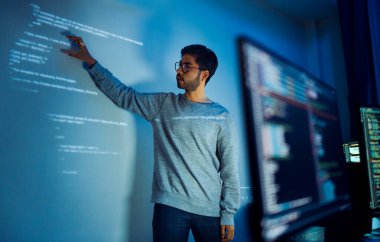 Hintli programcı, kod projeksiyonuyla ekranın önünde duruyor. Teknolojinin ve insan uzmanlığının yazılım geliştirme entegrasyonunu sunuyor.