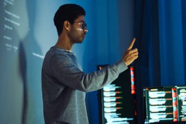 Hintli programcı, kod projeksiyonuyla ekranın önünde duruyor. Teknolojinin ve insan uzmanlığının yazılım geliştirme entegrasyonunu sunuyor.