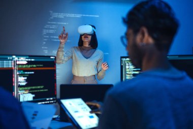 Geliştirici projeksiyondaki arayüz tasarımlarıyla etkileşime geçmek için bir VR kulaklığı kullanırken, meslektaşları ekranlarda sanal gerçeklik ve UI / UX tasarımının modern yazılım gelişimindeki entegrasyonunu tartışıp kodluyorlar..