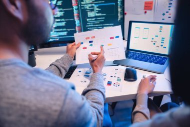 Asyalı yazılım geliştiricileri, modern ofiste gece yarısı kod ve uygulama diyagramlarını gösteren birden fazla ekran üzerinde çalışıyorlar.