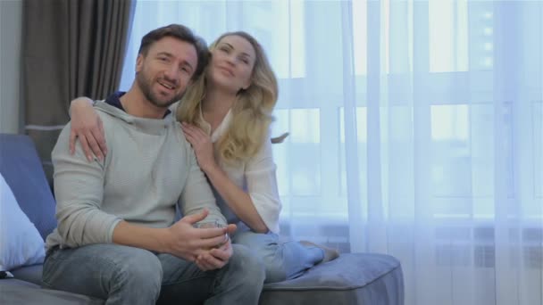 素敵なカップルは 自宅のソファに座っています かなり金髪女性のブルネットの後ろに座ってあごひげを生やした 魅力的なカップルのカジュアルな家庭の肖像画 — ストック動画