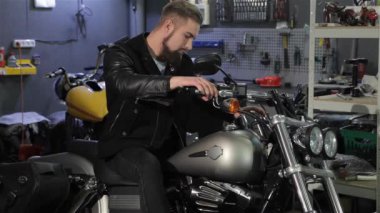 Genç erkek motorcu motosiklet Atölyesi'nde klasik helikopter poz. Serin sakallı adam kollarını göğsünde katlanır. Siyah deri ceket toching farklı parçaları üzerinde çekici beyaz adam