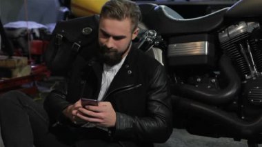 Erkek motorcu motosiklet atölyesinde telefonunu yakınındaki helikopter kullanarak. Motosiklet yakınındaki elinde smartphone ile gülen çekici beyaz adam. Genç adam motosiklet üzerinde eğilerek sakallı