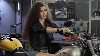 Genç kadın motorcu parmağını motosiklet üzerinde gösteriliyor. Çok kıvırcık kadın motosiklet inceleyerek siyah deri ceketli. Chopper motosiklet Atölyesi'nde onaylama çekici beyaz kız