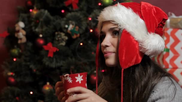 いくつかのホット飲料を飲んでサンタ帽子で若い白人女性 クリスマス ツリーの背景赤カップを保持している魅力的な女性 クリスマス近くの Copyspace を見てかなりブルネットの女性 — ストック動画