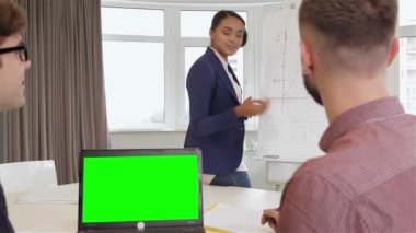 Güzel siyahi kadın erkek meslektaşlarına bazı projeler sunuyor. Çekici Afrikalı Amerikalı kız, tablonun yanında duruyor. Laptopun önünde oturan iki adam yeşil krom.