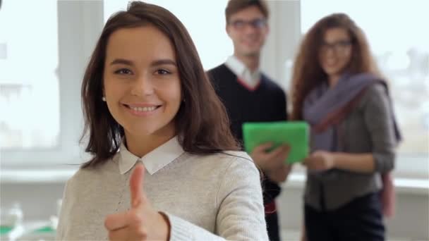 オフィスで良い品質をジェスチャーする笑顔のブルネットガール 親指を持ち上げた 白人少女の背景に立っている眼鏡の男性と女性 カーリー ブルネット タブレット — ストック動画