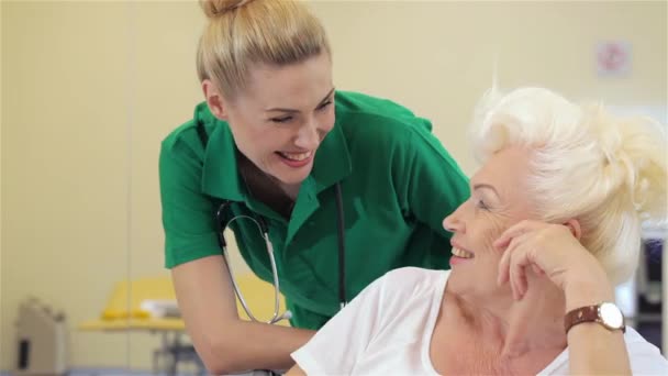有吸引力的年轻护士微笑岁女病人在医院 白种人金发碧眼的护士站高级女人的背后 漂亮的护士在绿军装和老妇在镜头前微笑 — 图库视频影像