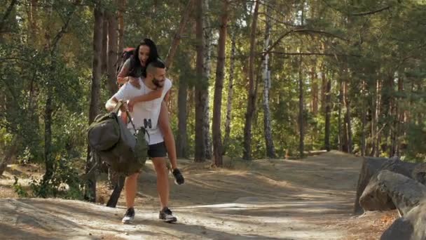 彼女の彼氏の背中に乗って女性ハイカーのスローモーション 森で楽しんで幸せな若いカップル 彼氏の頬をキスかなりブルネットの少女 — ストック動画