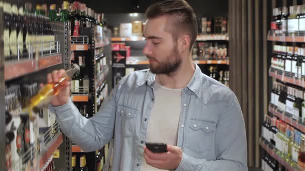 年轻人在超市里选择饮料 高加索人在超级市场用他的智能手机留着胡子的男人穿着蓝色衬衫和白色T恤 看着他的手机 — 图库视频影像