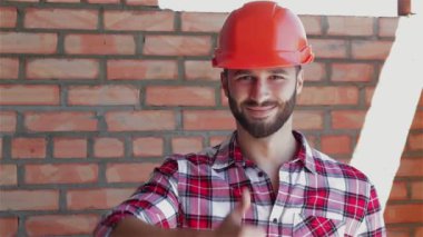 Erkek inşaatçı inşaat aşamasındaki binada başparmağını gösteriyor. Turuncu sert şapkalı sakallı adam tuğla duvar arka plan aleyhine duruyor. Genç inşaat işçisi önünde elini kaldırıyor