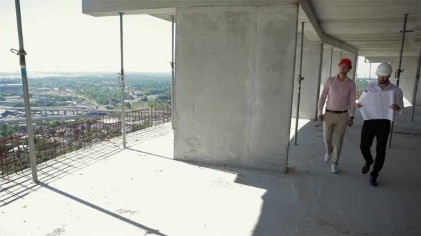 两名男性建筑工人在施工中穿过大楼的一层 建筑专家来到地板的边缘 胡子建筑师手里拿着计划 — 图库视频影像