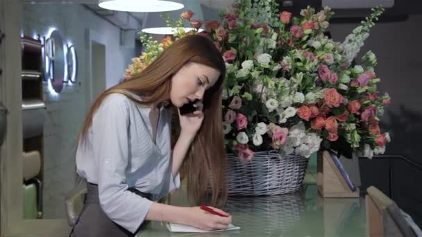 フラワー ショップに電話で話している女性の花屋の側面図です 魅力的な若い女性の花の背景に赤のペンで書きます 花の注文を引き継ぐかなり白人の女性 — ストック動画