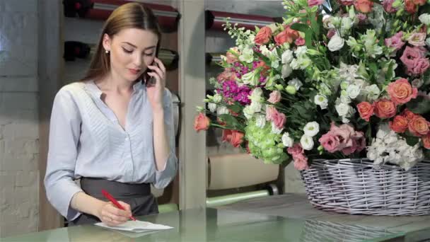 女性の花屋フラワー ショップで大きな花束の注文を受け入れます かなり白人の女性は彼女のノートの順序を書いている間彼女の耳の近くの彼女の携帯電話を保持しています 魅力的な若い女性 — ストック動画