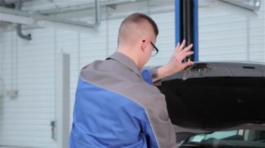 Araba hood kapanış mavi üniformalı genç makinist. Sarışın beyaz adam ellerini bir bezle silerek. Gözlük kamera için gülümseyen otomotiv uzmanı