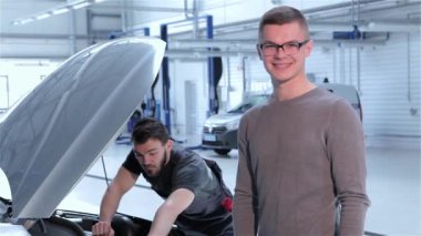 Genç istemci araba servisi onaylama. Araba Servisi parmağını yukarı kaldırarak gözlük sarışın adam. Beyaz adam arka plan çalışma mekanik üzerinde duran gri Kazak giymiş