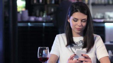 Telefonunu restoranda bakarak çekici esmer kadın. Tablo kırmızı şarap bardak alarak güzel kız. Güzel beyaz kız onun smartphone kullanırken bir yudum şarap yapma