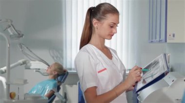 Kadın dişçinin dişçi dolabında bir şey işaretlediğini gösteren bir yan görüş. Tıbbi üniforma giymiş, elinde mavi kalemle beyaz bir kadın. Genç diş uzmanı.
