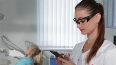 Genç diş uzman diş hekimi kabine, onun akıllı telefon dokunmatik ekran üzerinde yazarak. Koruyucu gözlük kamera için gülümseyen çekici beyaz kadın. Onu koyarak güzel brownhaired kadın