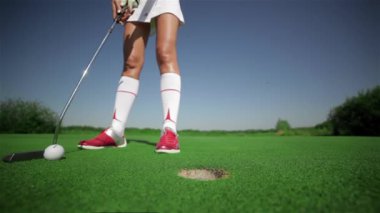 Dişi golfçü deliklerini bir vuruşla kapat. Kırmızı golf ayakkabılı genç bir kadın topa hafifçe vuruyor. Beyazlı kadın golf sopasını elleriyle tutuyor.