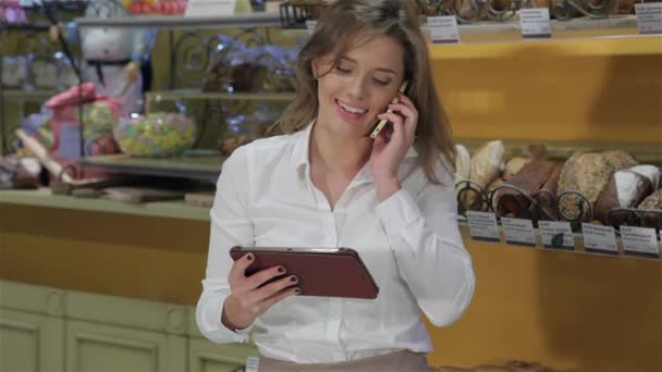ペストリーショップで電話で話しているかわいい女性 彼女の手にデジタルタブレットを保持している魅力的な白人女性 デジタルタブレット画面を見て若い女性の売り手 — ストック動画