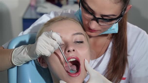 キャビネットの歯科で患者の上顎中切歯を調べる女性歯科スペシャ リストのクローズ アップ 患者さんのあごを保持保護メガネの若い Brownhaired 歯科医 かなり金髪女性 — ストック動画