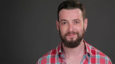 Sırıtarak modern birini kapatın. Kameraya bakarak Küpeli esmer hippi. Sakallı adam koyu gri arka plan üzerinde izole kareli gömlek giymiş Küpeli