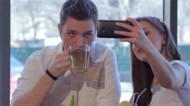 Çekici kız yakışıklı erkek arkadaşıyla selfie çekiyor. Beyaz adam çayını içerken güzel kızı da kendi fotoğrafını çekiyor. Elinde siyah bir akıllı telefon tutan gülümseyen bir kadın. Onun ve onun önünde.