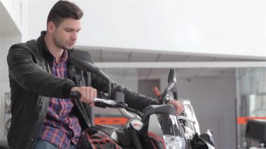 Siyah motosiklet üzerinde oturan ileri genç satıcı yakından. Beyaz adam dirseklerinden motosikletin benzin deposuna yaslanmış. Kameraya bakan yeni motosiklet üzerinde hafif sakallı esmer