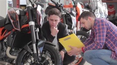 Motosiklet salonunda spor motosiklete bakan iki genç adam. Danışman siyah spor motosiklet hakkında tüketici ile konuşurken damalı gömlek giymiş. Tüketici siyah ceket ve beyaz giymiş