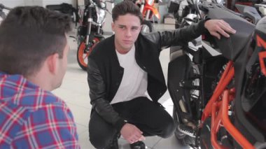 Müşteriye spor turuncu motosiklet gösteren satıcı. Motosikletin koltuğuna ve çerçevesine dokunan genç adam. Danışman çerçeveişaret motosiklet özellikleri hakkında söylüyorum