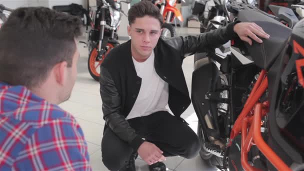顧客にスポーツオレンジのオートバイを示すディーラー バイクの座席とフレームに触れる若い男 フレームを指すバイクの特徴を語るコンサルタント — ストック動画