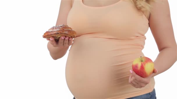 リンゴとケーキの重量を量る妊娠中の女性は クローズアップ 妊娠中の女性は 片手にリンゴを持ち 別の手でケーキを持っています 妊娠中のリンゴとケーキを白い背景の上に隔離 — ストック動画