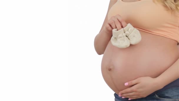孕妇把婴儿战利品放在肚子上 怀孕肚子上白色婴儿靴的特写 怀孕的白种人抚摸着她的肚子在白色背景 — 图库视频影像
