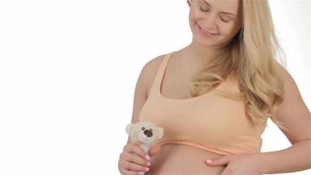白い背景にテディベアによって手を振ってかなり妊娠中の女性 妊娠中のブロンドは 別の手でおもちゃを保持しながら 彼女の腹をなでる おもちゃを演奏妊娠中の白人女性のスタジオショット — ストック動画