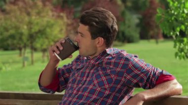 Bankta oturan ve dışarıda kahve içen bir erkek portresi. Bir şehir parkında öğrenciler veya gençlerin geri kalanı. Sağlıklı eğlence ve eğlence kavramı.