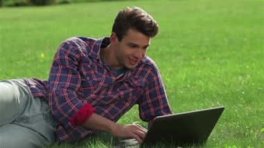 Yakışıklı genç adam parkta çimenlerin üzerinde yatıyor ve dizüstü bilgisayarla çalışıyor. Bir şehir parkında öğrenciler veya gençlerin geri kalanı. Sağlıklı eğlence ve eğlence kavramı.
