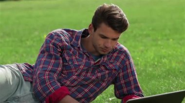 Genç yakışıklı adam parkta çimlere uzanıyor ve dizüstü bilgisayar kullanıyor. Öğrencilerin geri kalanı ya da şehir parkındaki gençler. Sağlıklı eğlence ve eğlence kavramı..