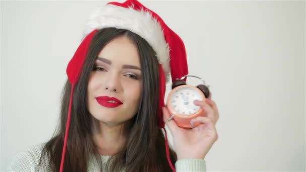 戴着圣诞礼帽的漂亮姑娘摆着钟 表示新年还将持续5分钟 在白色的背景上微笑快乐的女孩 漂亮的女人 戴着圣诞礼帽的女人 — 图库视频影像