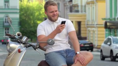 Yakışıklı sakallı genç adam scooter ile smartphone üzerinde oturuyor. Adam scooter üzerinde oturan ve smartphone vasıl arıyorsunuz