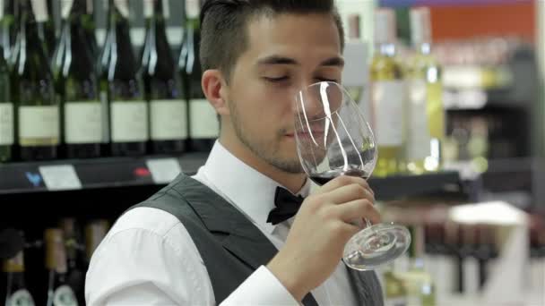 自信和有经验的品酒师 自信的年轻男子戴着领结控股葡萄酒玻璃用红酒和看其纹理与葡萄酒架在背景中 — 图库视频影像